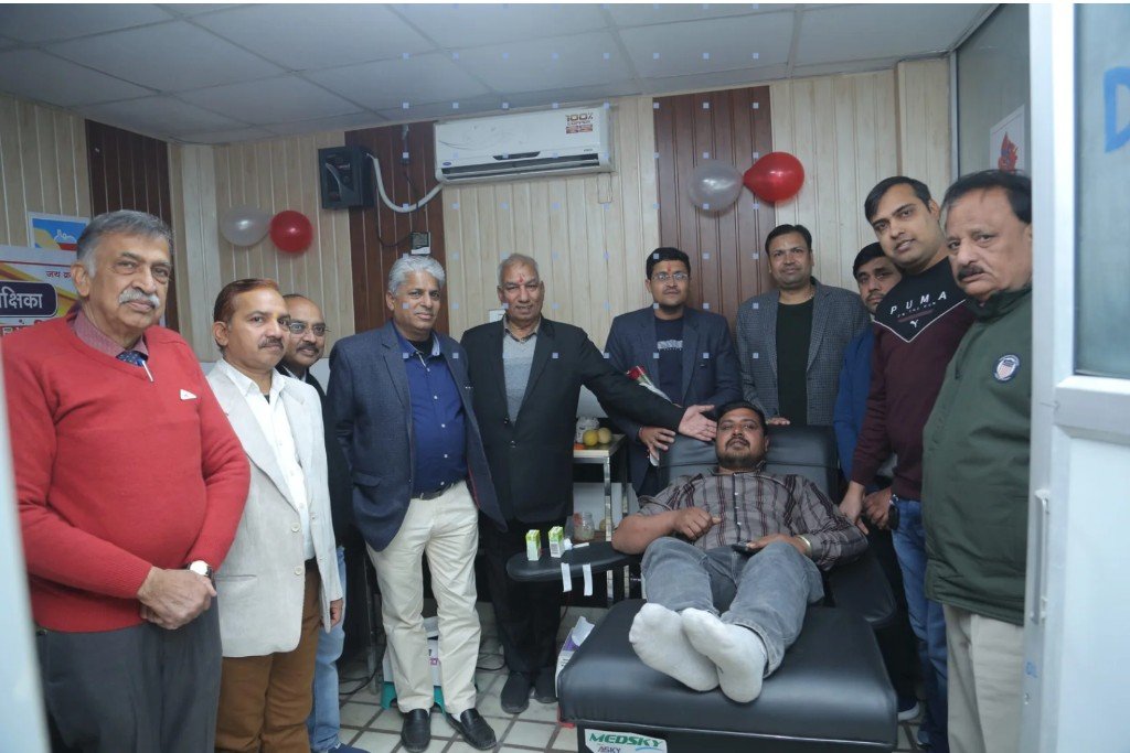Indoor blood donation event: a donor reclines in a medical donation chair as a group poses nearby; room with wood panelling and festive balloons — Manav Seva Sangam Hospital, Tohana